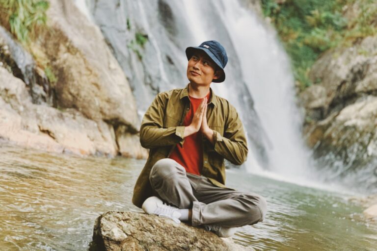 Spiritual Healing man in brown jacket sitting on rock near water falls during daytime